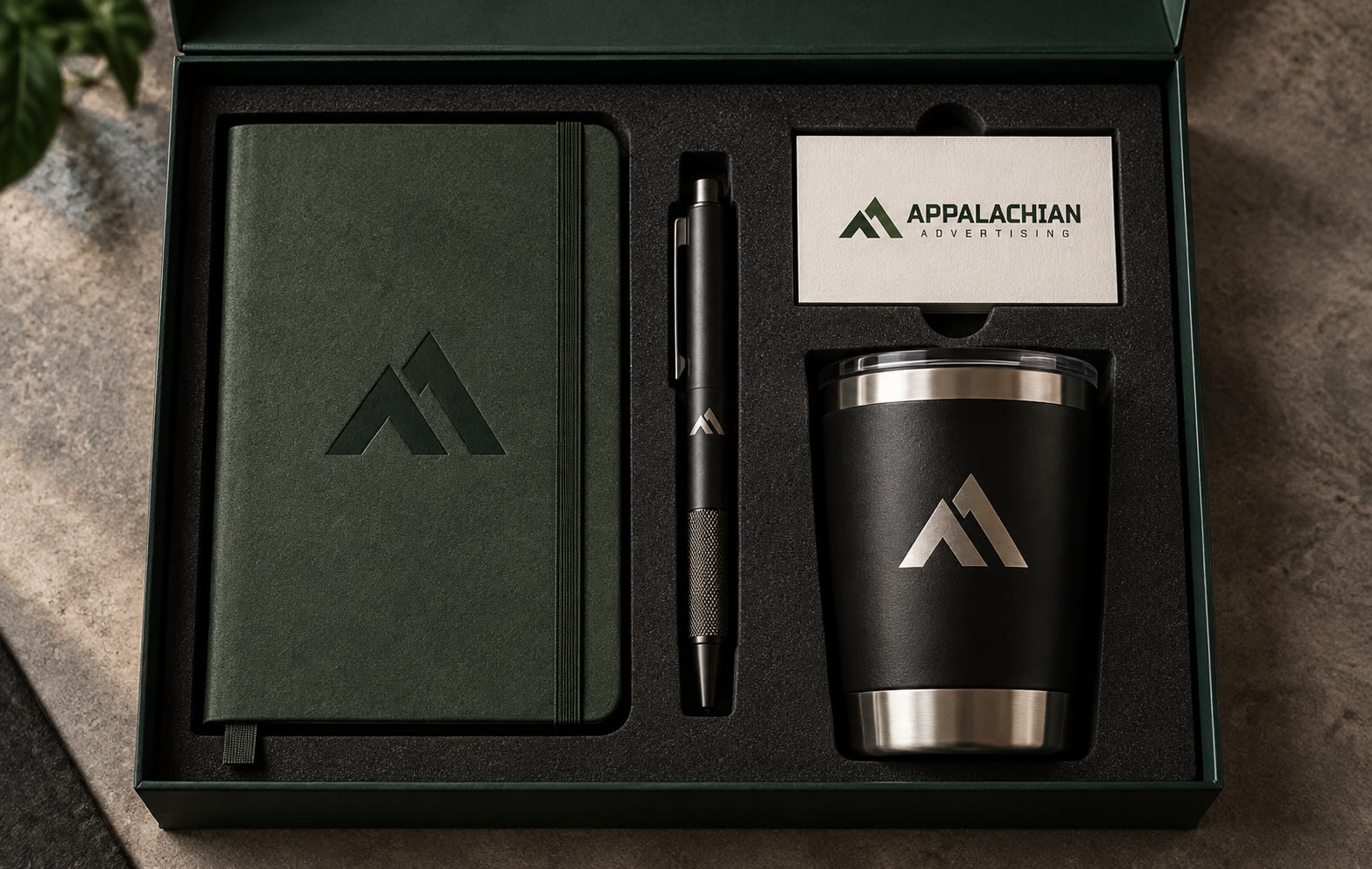 A Spruce gift box holding a debossed notebook, a pen, a business card with the horizontal lockup, and a black tumbler with the emblem etched in steel.